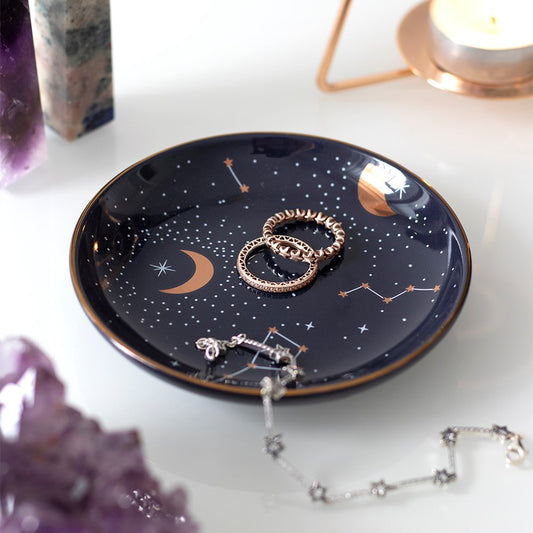 Dark blue ceramic trinket dish with a metallic gold constellation design. The dish is holding some pieces of jewellery and placed on a dressing table, surrounded by crystals.