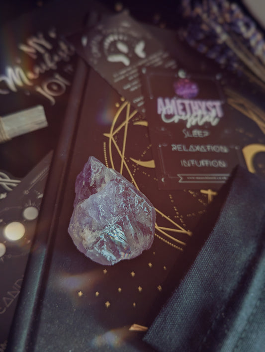 A chunk of purple raw amethyst rests upon a black journal embossed with a mystical gold design, surrounded by crystals and sprigs of lavender. In the background we can see a crystal care card containing properties of amethyst crystals and a black cotton drawstring bag.